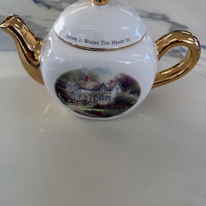 May include: A white ceramic teapot featuring a gold-colored handle, spout, and lid knob. The lid is inscribed with the text "HOME IS WHERE THE HEART IS" in black lettering. A colorful illustration of a house with a garden is on the front.