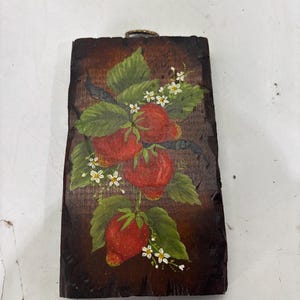 May include: A rectangular wooden plaque with a dark brown stained finish and rough, carved edges. The surface features a hand-painted illustration of three bright red strawberries with green leaves and small white flowers, including a metal hanger.