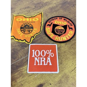 May include: Three embroidered patches: an orange Ohio-shaped patch with black text reading OHIO DIVISION OF WILDLIFE SAFE HUNTER, a circular orange patch with black deer silhouettes and text, and a square orange patch with white text reading 100% NRA.