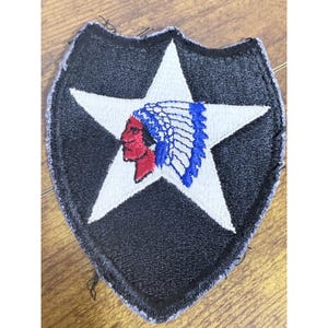 Puede incluir: Un parche bordado en forma de escudo con fondo negro y una gran estrella blanca en el centro. Dentro de la estrella hay una ilustración de perfil de una persona con piel roja que lleva un tocado de plumas azul y blanco.