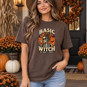 May include: A dark brown short-sleeve t-shirt featuring a graphic of a skeleton wearing sunglasses and a hair bow, holding a pumpkin-themed cup. The text reads BASIC WITCH BUT MAKE IT SPOOKY, surrounded by an orange circle, bats, and stars.