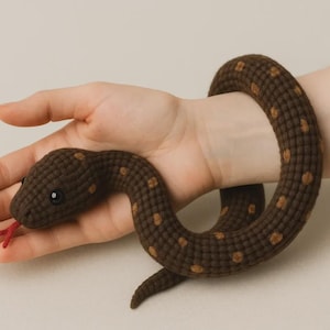 Può includere: Un serpente giocattolo all'uncinetto marrone scuro con macchie marrone chiaro, un occhio a perla nera e una lingua in filo rosso. Il serpente è avvolto in una forma circolare, caratterizzato da una struttura in filo intrecciato su tutto il corpo.