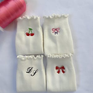 May include: Four white ankle socks with ruffled cuffs, each featuring a unique embroidered design: a pair of red cherries, a pink bow above the letter M, the cursive initials DS in black, and a red bow. Two spools of thread are visible.