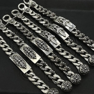 Puede incluir: Una colección de cinco pulseras de metal plateado con cadenas de eslabones gruesos. Cada pulsera incluye una placa central rectangular grabada con un motivo de cruz floral o daga, terminada con un cierre circular en un extremo.