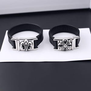 Puede incluir: Dos pulseras de cuero negro con cierres de metal plateado. La pulsera de la izquierda tiene un emblema de flor de lis en el cierre, mientras que la pulsera de la derecha presenta un diseño floral circular de varios pétalos en su cierre metálico.