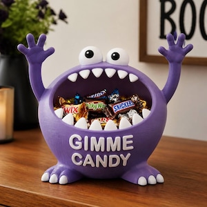May include: A purple monster-shaped candy bowl with two large white eyes, raised arms, and white teeth lining a wide open mouth. The text GIMME CANDY is embossed in white on the front. The bowl is filled with various assorted chocolate candy bars.