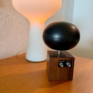 May include: A black spherical speaker mounted on a square wooden base with a metal rod. The base features a black control panel labelled LLOYDS SOLID STATE YORICK with two silver knobs and a small dial for volume and tuning adjustments.