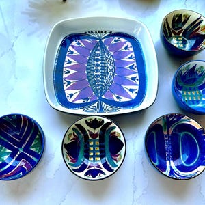 Puede incluir: Un plato de servicio cuadrado de cerámica y cinco cuencos redondos con un vibrante patrón de inspiración floral en azul, morado y blanco. El conjunto incluye una fuente cuadrada grande y cinco cuencos más pequeños, decorados con motivos botánicos simétricos.