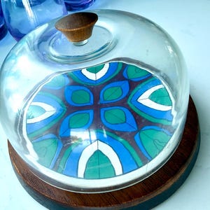 May include: A glass dome food cover with a dark brown wooden base and a matching wooden knob handle. The base features a circular inlay with a repeating geometric pattern in shades of blue, green, white, and dark brown, visible through the clear glass.