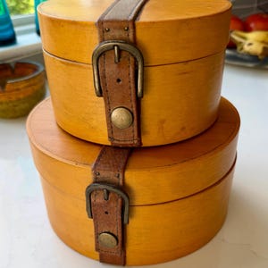 May include: Two round, light brown wooden storage boxes stacked vertically. Each box features a brown leather strap that wraps around the centre, secured with a brass-toned metal buckle and a matching circular metal stud at the base of the strap.