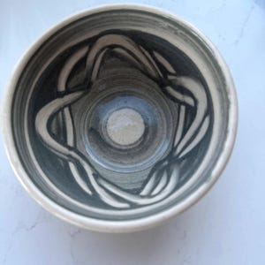 Könnte beinhalten: Eine Keramikschale mit kreisförmiger Öffnung und einem glatten, hellen Rand. Das Innere zeigt ein handgemaltes, dunkelgraues, wirbelndes seilartiges Muster, das die Basis umgibt und sich von der hellgrauen und weißen glasierten Oberfläche abhebt.