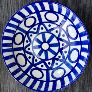 May include: A round ceramic bowl featuring a white base with a hand-painted cobalt blue pattern. The design includes a central starburst, a ring of circular shapes, and a wide outer border composed of alternating blue and white vertical stripes.