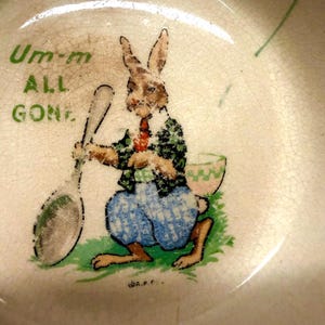 May include: A cream-colored ceramic bowl with a crackled glaze finish featuring a graphic of a brown rabbit wearing a green patterned jacket and blue trousers. The rabbit holds a large spoon, and the text reads Um-m ALL GONE in green letters.