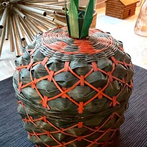 Puede incluir: Un recipiente redondo con tapa en forma de piña, construido con tiras tejidas de color verde oscuro y acentuado con un cordón naranja brillante en un patrón entrecruzado. La tapa presenta un pequeño adorno de hoja verde en el centro.