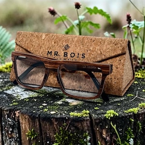 Peut inclure: Lunettes rectangulaires avec une monture en bois brun et des verres transparents, posées contre un étui triangulaire en liège. L'étui porte l'inscription MR. BOIS imprimée en noir sur la face avant, située au-dessus des lunettes.