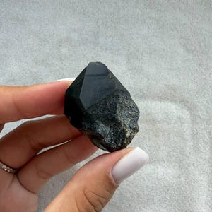 以下が含まれることがあります： 滑らかでファセット加工された頂点と、粗く質感のある基部を持つ、原石の黒い結晶標本。この石は深く不透明な黒色をしており、磨かれた幾何学的な頂点からギザギザした底部へと変化する、自然で不規則な形状をしています。