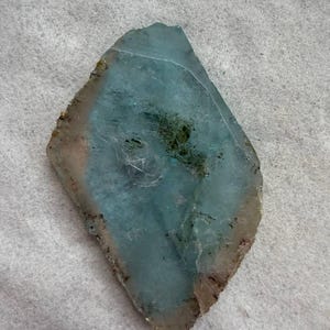 May include: A flat, irregularly shaped slice of translucent blue-green mineral with a rough, light-coloured outer edge. The centre displays a cloudy, semi-transparent texture with darker green inclusions and fine, thread-like white internal patterns.