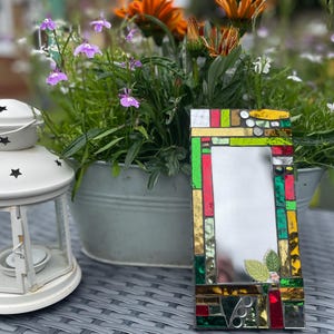 May include: A rectangular mirror with a mosaic frame made of colorful glass tiles. The frame features shades of red, green, yellow, and brown. The mirror is surrounded by a variety of flowers in a pot.