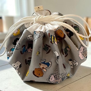 May include: A gray fabric drawstring pouch featuring a repeating pattern of Peanuts characters including Snoopy, Charlie Brown, and Lucy. The bag has a white interior lining, white cord drawstrings, and a small beige tag labeled handmade with.