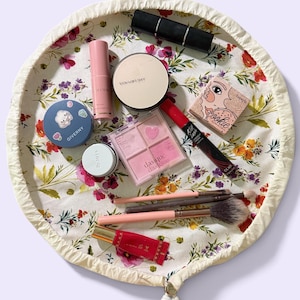 May include: A round, drawstring makeup bag featuring a floral pattern with red, yellow, and green flowers on a white background. The bag contains various cosmetics, including a pink eyeshadow palette, a black mascara, brushes, and compacts.