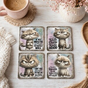 May include: A set of four square coasters featuring illustrations of grumpy cats with text. The top left reads IF MY MOUTH DOESN'T SAY IT MY FACE DEFINITELY WILL. Top right reads Kinda Classy Kinda HOOD. Bottom left reads I DON'T HAVE THE ENERGY TO PRETEND I LIKE YOU TODAY. Bottom right reads I'm not Responsible for what my face does when you talk.