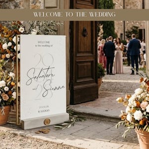 Puede incluir: Un letrero de bienvenida de boda rectangular blanco con texto en caligrafía negra que dice Salvatore and Sienna, la fecha 20.04.28 y el texto IL LUOGO. El letrero está en un marco de metal dorado sobre una base de hormigón con un sello de cera dorado.