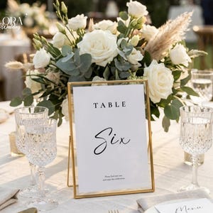 Puede incluir: Un letrero de mesa rectangular vertical con un marco fino de metal dorado que muestra el texto TABLE SIX en tinta negra. El letrero está colocado frente a un gran arreglo floral con rosas blancas, hojas de eucalipto y hierba de la pampa seca.