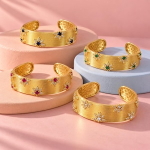 Peut inclure: Quatre bracelets manchette dorés avec une surface texturée et striée. Chaque bracelet présente des motifs en forme d'étoile espacés, ornés de petites pierres claires et d'une pierre centrale colorée bleue, verte, rouge ou transparente.