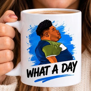 May include: A white ceramic mug featuring a graphic illustration of a person with dark curly hair wearing a blue and olive green shirt against a blue paint splatter background, with the black text "WHAT A DAY" printed clearly below the figure.