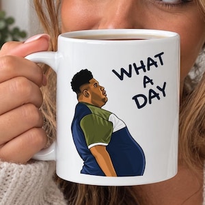 May include: A white ceramic mug featuring a graphic illustration of a person with a dark complexion and short hair, wearing a blue and olive green shirt. The text "WHAT A DAY" is printed in dark blue capital letters next to the illustration.