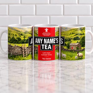 May include: A white ceramic mug featuring a landscape of green hills, a stone wall, and sheep. The centre displays a red label with the text "ANY NAME'S TEA" and "Let's have a proper brew" above "TEA BAGS" and a small white emblem at the top.