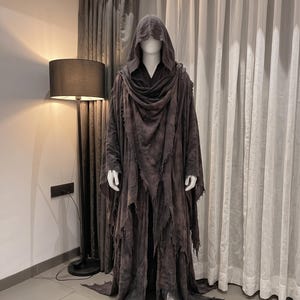 Peut inclure: Une robe à capuche gris foncé pleine longueur présentant un ourlet effiloché et usé, avec des couches de tissu drapées autour des épaules et de la poitrine. Le vêtement possède de longues manches larges et une capuche profonde en tissu texturé.