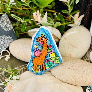 May include: Hand-painted rock art featuring a cartoon giraffe with orange and brown spots, surrounded by colorful flowers and leaves on a blue background.