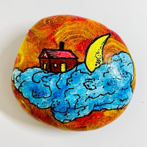 Puede incluir: Una piedra pintada a mano con una casa roja sobre una nube azul, una luna creciente amarilla en el fondo y un fondo rojo, amarillo y naranja.