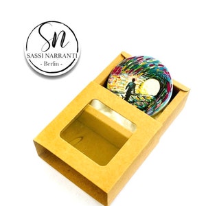 Könnte beinhalten: Ein rundes, abstraktes, farbenfrohes Gemälde einer Person, die auf einem Weg mit einem weißen Kreis im Hintergrund geht. Das Gemälde befindet sich in einer braunen Pappbox mit einem durchsichtigen Fenster.