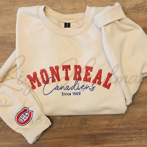 Op de afbeelding: Een crèmekleurig sweatshirt met ronde hals en rode geborduurde tekst MONTREAL, met daaronder de blauwe cursieve tekst Canadiens en de blauwe tekst Since 1909. Een rood, wit en blauw logo is geborduurd op de linker mouwboord.
