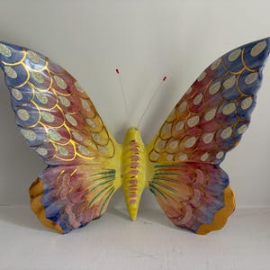 May include: A decorative butterfly wall hanging featuring a yellow central body and two large wings with a scalloped pattern. The wings display a gradient of blue, pink, and orange hues, accented with gold outlines and silver glitter details.