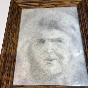May include: A framed charcoal portrait drawing of a person with a soft, hazy appearance, encased in a rectangular wooden frame with a prominent dark brown wood grain pattern and a beveled edge design surrounding the artwork.