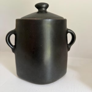 May include: A matte black ceramic canister with a rounded square base, two side handles, and a matching lid featuring a small knob handle. The container has a smooth, dark finish and a vertical, slightly tapered cylindrical body shape.