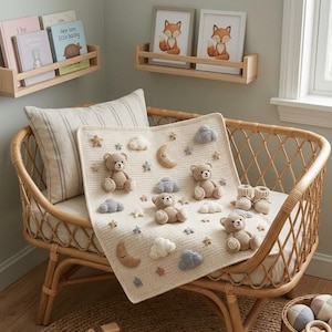 May include: A cream-colored knit baby blanket featuring attached three-dimensional crochet teddy bears, crescent moons, clouds, and stars in shades of beige, gray, and white, accompanied by a pair of matching small knit booties resting on the fabric.