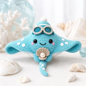 Op de afbeelding: Een lichtblauw gehaakt pijlstaartrog knuffeltje met witte stippen, zwarte veiligheidsoogjes, roze wangen en een kleine beige schelp in de vinnen met een witte parel. De rog draagt een blauw-witte gehaakte badmuts en een duikbril.