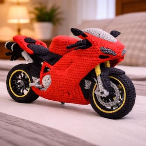 May include: A handcrafted crochet motorcycle toy featuring a vibrant red body with black accents, a grey windshield, and black wheels with yellow rims. The detailed fiber art piece displays a sport bike silhouette with visible handlebars and seat.