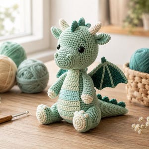 May include: A crocheted dragon toy in shades of sage green and cream with dark green wings, black safety eyes, and small horns. The dragon features a textured stitch pattern, a long tail with spikes, and a light-colored belly panel.
