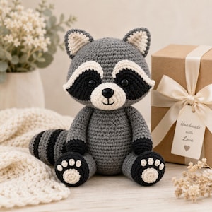 Puede incluir: Un peluche de mapache tejido a ganchillo con cuerpo gris, parches negros en los ojos, hocico blanco y cola a rayas. El mapache tiene patas negras con detalles blancos y una pequeña nariz negra. Una etiqueta de regalo dice Handmade with Love.