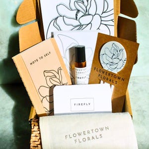 May include: A cardboard gift box containing a tan notebook labeled Note to Self, a small amber glass roller bottle labeled Flowertown Florals Firefly, a white card labeled Firefly, a brown card with a floral sticker, and a white fabric pouch.