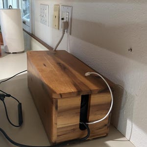 May include: A rectangular wooden cable management box with a natural brown finish. The box features a removable lid and a vertical slot on the front panel for routing cords. A white charging cable extends from the top, and a black cord exits the front.