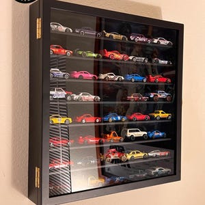 May include: A black rectangular wall-mounted display case with a glass door and gold-colored hinges, containing multiple shelves filled with various miniature toy cars in diverse colors. The logo Woodland Crate Co is visible in the top corner.