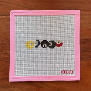 May include: A square needlepoint canvas featuring a pink fabric border. The center displays a row of five billiard balls in yellow, blue, black, and red, with the number eight on the black ball. The text XOXO is stitched in red in the bottom corner.