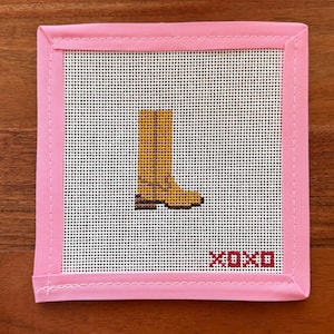 May include: A square needlepoint canvas featuring a yellow boot design in the center and the text XOXO in red at the bottom right corner. The canvas is finished with a light pink fabric border stitched around all four edges of the square piece.