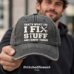May include: A charcoal grey, acid-washed baseball cap featuring white embroidered text that reads: THAT'S WHAT I DO I FIX STUFF AND I KNOW THINGS, with a crossed wrench and hammer icon integrated into the design on the front panel.